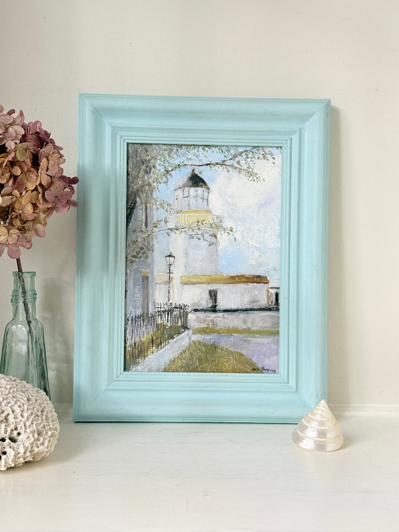 A framed art print depicting a lighthouse scene, styled on a white surface with a glass vase of dried flowers and a small seashell against a cream wall.
