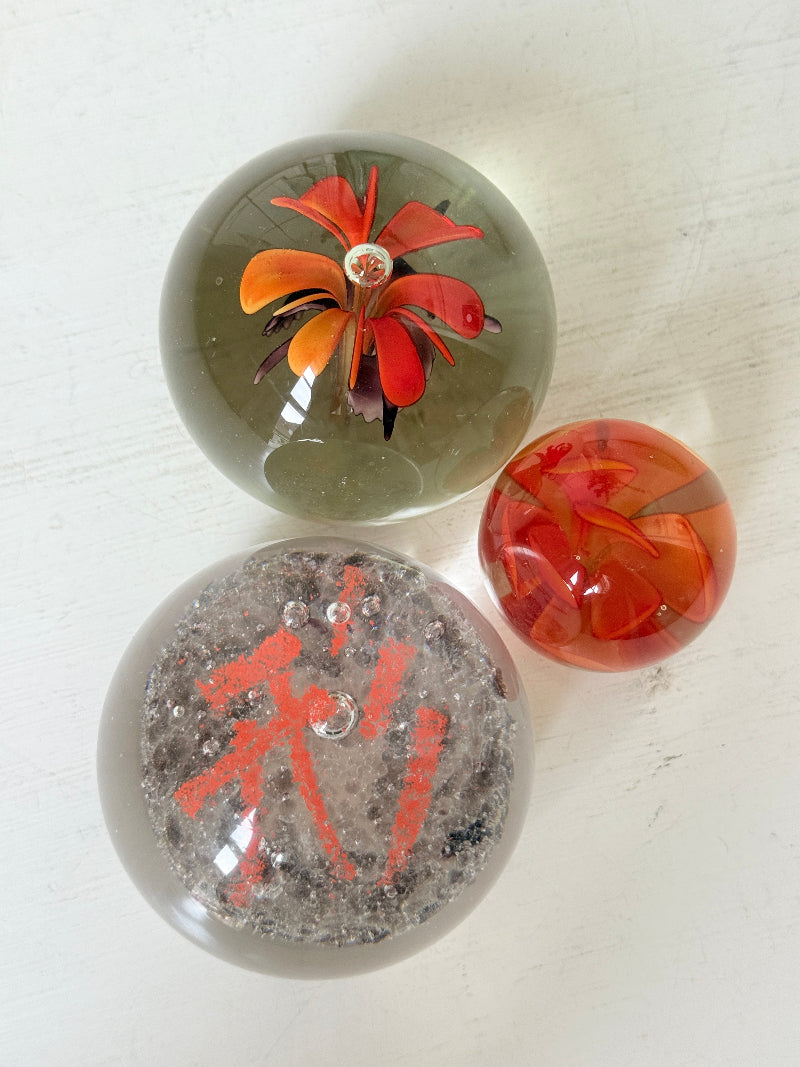 Trio of orange flower glass paperweights with green stems and controlled bubbles, photographed on a white surface