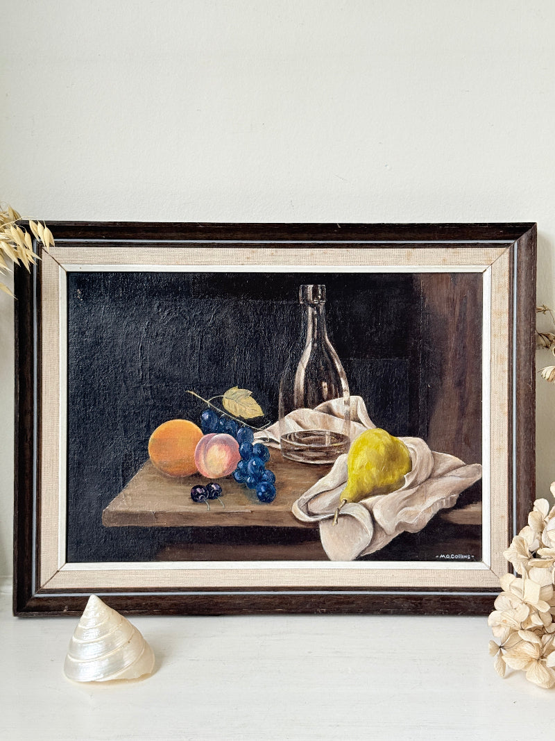 A framed still life oil painting depicting fruit, grapes, a pear, and a glass bottle on a tablecloth against a dark background, presented in a dark wood frame on a white surface with decorative dried flowers and a shell.