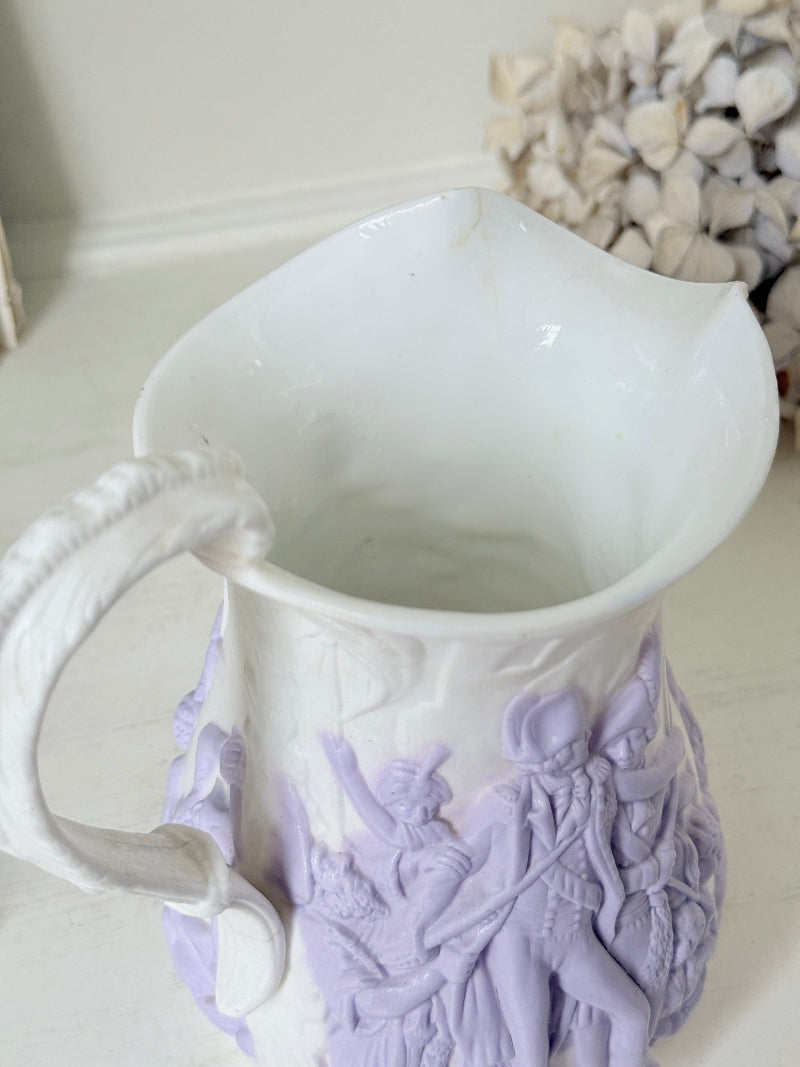A white ceramic pitcher with a light purple relief decoration depicting classical figures, shown on a light-colored surface with a blurred background.