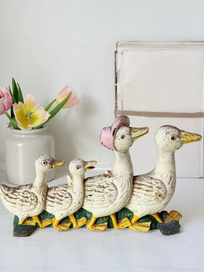 A decorative family of four duck figurine set, featuring distressed cream-colored bodies with brown speckles, yellow beaks and feet, and a pink hat on the adult duck. The figurines are displayed on a white surface next to a white vase with pink and yellow tulips.