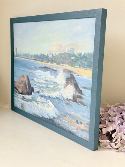 Original oil painting of waves crashing against rocks, signed seascape in slate blue wooden frame on white surface

