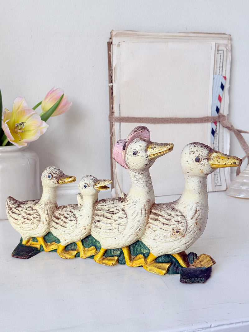 A decorative cast-iron duck family figurine set on a white tabletop, featuring a large duck with a pink hat and three smaller ducks behind it, with yellow beaks and feet. A vase with tulips and a rustic framed print are in the background.