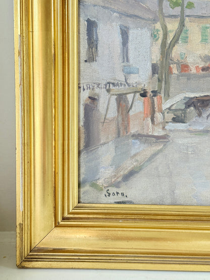 A close-up of a framed painting showing part of a street scene with buildings, figures, and a tree, enclosed in a wide ornate gold frame. The painting has a soft impressionistic style, and the artist&