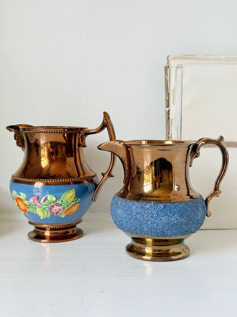 Two vintage ceramic pitchers with metallic copper-colored handles and bases, one decorated with a colorful floral band and the other with a speckled blue lower section, displayed on a white surface against a white wall.