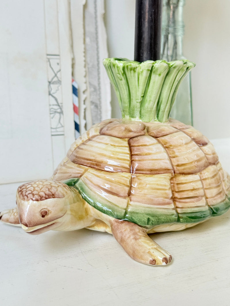 A ceramic turtle-shaped candle holder with a green leafy top, shown on a light surface with a black candle inserted.