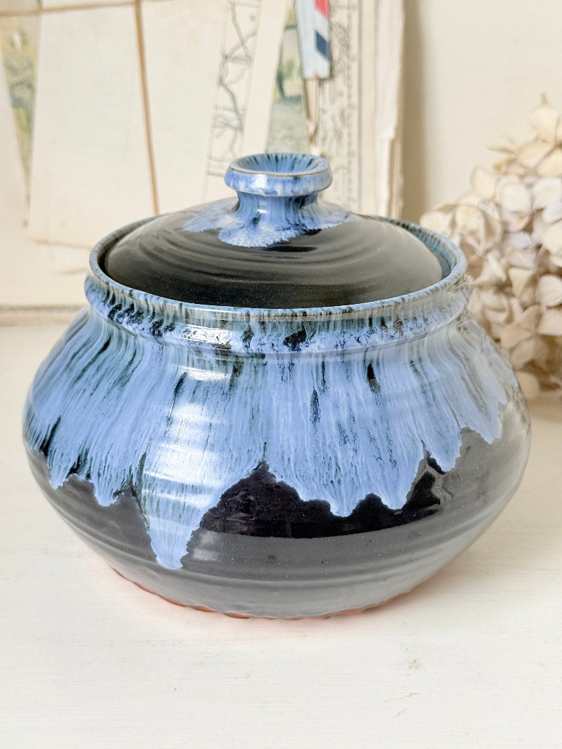 A handmade ceramic lidded jar with a glossy glaze, featuring a dark black base with a dripping light blue glaze effect, set against a softly blurred background.