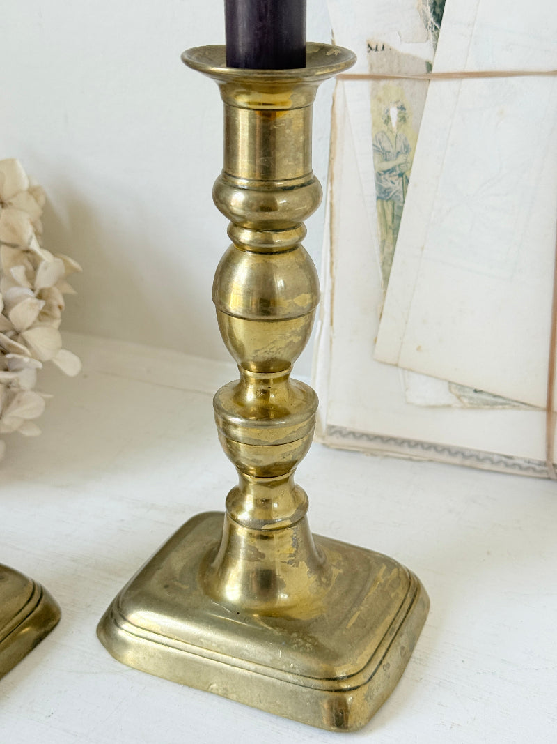 Pair of turned brass candlesticks with square stepped bases and black taper candles on a white surface, styled with dried flowers