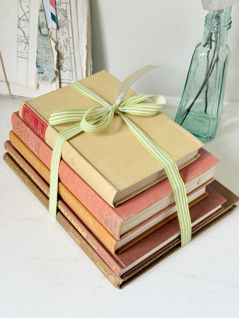 A stack of vintage hardcover books tied with a light green and white striped ribbon, set on a white surface with a glass bottle in the background.