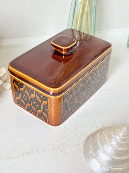A rectangular ceramic butter dish with a glossy brown glazed lid and base, featuring a dark brown geometric pattern on the sides, set on a white wooden surface.