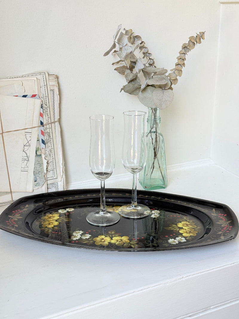 A black oval decorative tray with yellow and white floral patterns, holding two clear champagne flutes, set on a white mantelpiece or shelf next to a glass vase with dried eucalyptus and a stack of vintage letters.