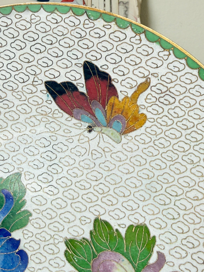Close-up of a decorative plate featuring a colorful butterfly motif in red, yellow, blue, and black on a white background with a repeating gold geometric pattern.