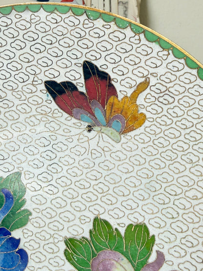 Close-up of a decorative plate featuring a colorful butterfly motif in red, yellow, blue, and black on a white background with a repeating gold geometric pattern.