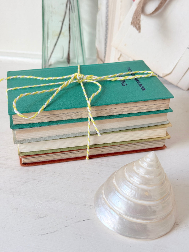 A stack of vintage hardcover books tied with yellow-and-white string, placed on a white wooden surface next to a clear seashell. A glass bottle and a cream fabric bag are blurred in the background.
