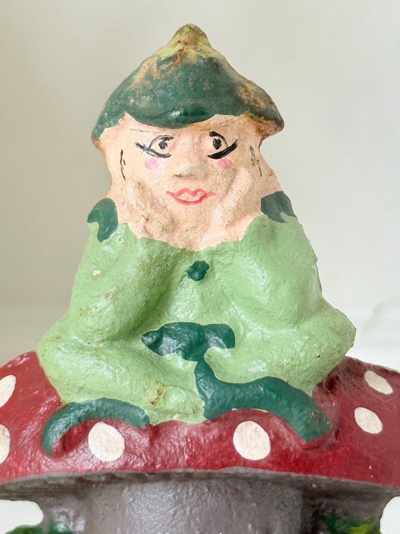 A close-up of a hand-painted gnome figurine sitting on a red mushroom with white polka dots. The gnome has a green hat and coat, with a weathered, rustic finish.