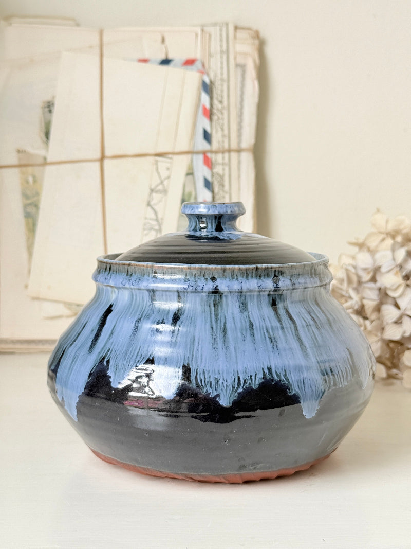 A handmade ceramic lidded jar with a glossy glaze, featuring a dark black base with light blue dripping glaze details and a terracotta-colored rim, set against a softly blurred background of vintage letters and dried flowers.