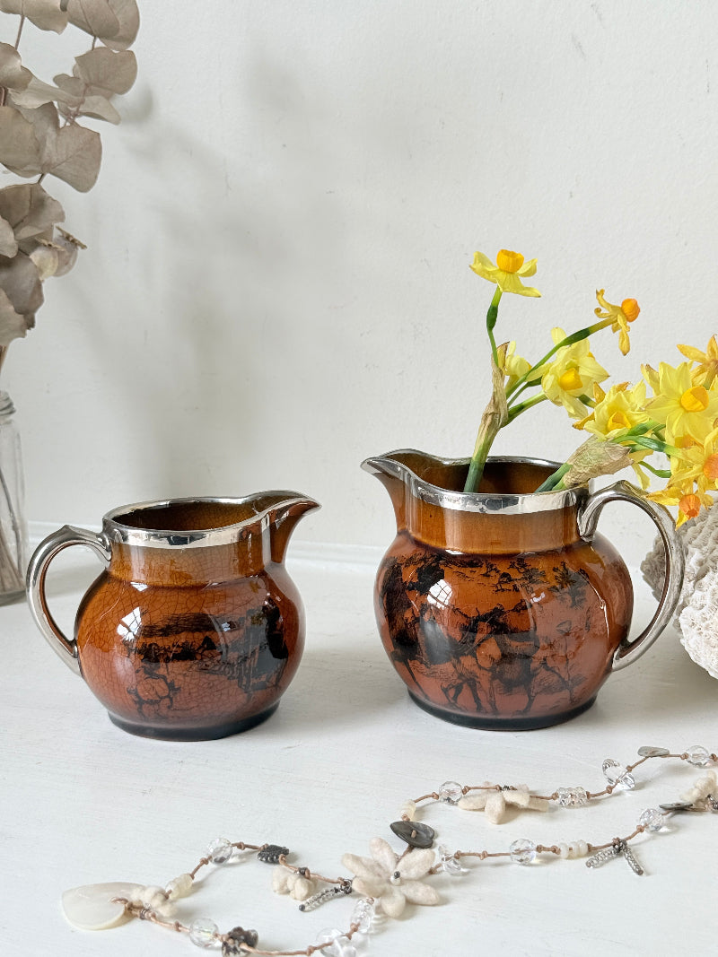 Pair of antique Ridgways stoneware creamer jugs photographed on a white surface in amber treacle glaze with black coaching scenes and silver lustre rims