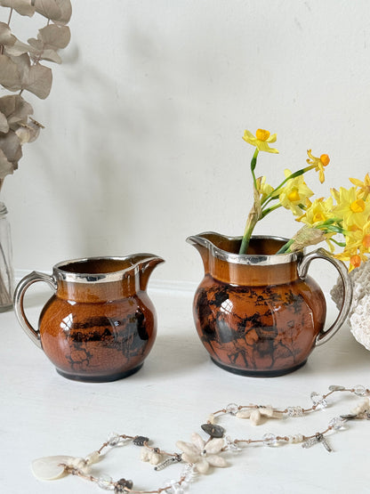 Pair of antique Ridgways stoneware creamer jugs photographed on a white surface in amber treacle glaze with black coaching scenes and silver lustre rims