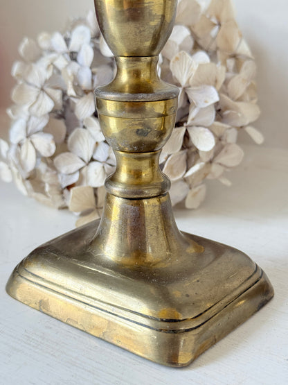 Pair of turned brass candlesticks with square stepped bases and black taper candles on a white surface, styled with dried flowers