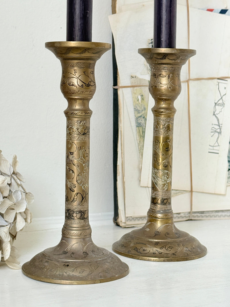Pair of tall Indian engraved brass candlesticks in the Benares style with floral detailing and black taper candles on a white surface