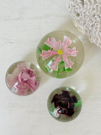 Trio of vintage glass paperweights with hand worked pink and plum floral detail, clear glass domes and green leaf decoration
