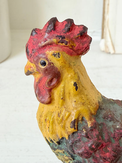 A close-up of a vintage cast iron rooster figurine with a distressed yellow body, red comb and wattle, and dark green and red wing feathers, set against a softly blurred white background.