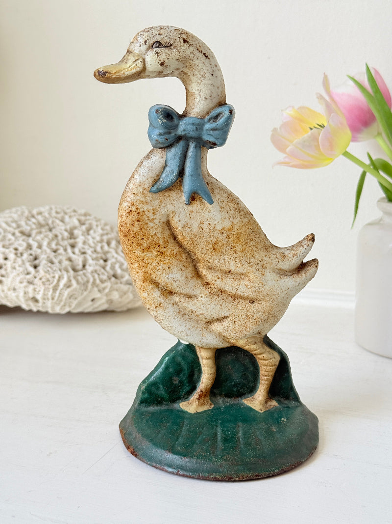 A vintage ceramic duck figurine with a speckled cream and brown glaze, wearing a blue bow tie and standing on a dark green base, displayed on a white surface with decorative background elements.