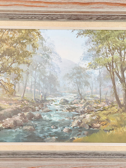 A framed landscape painting depicting a scenic river flowing through a wooded valley, with rocks in the water, trees, and distant mountains. The artwork is presented in a light wood frame.