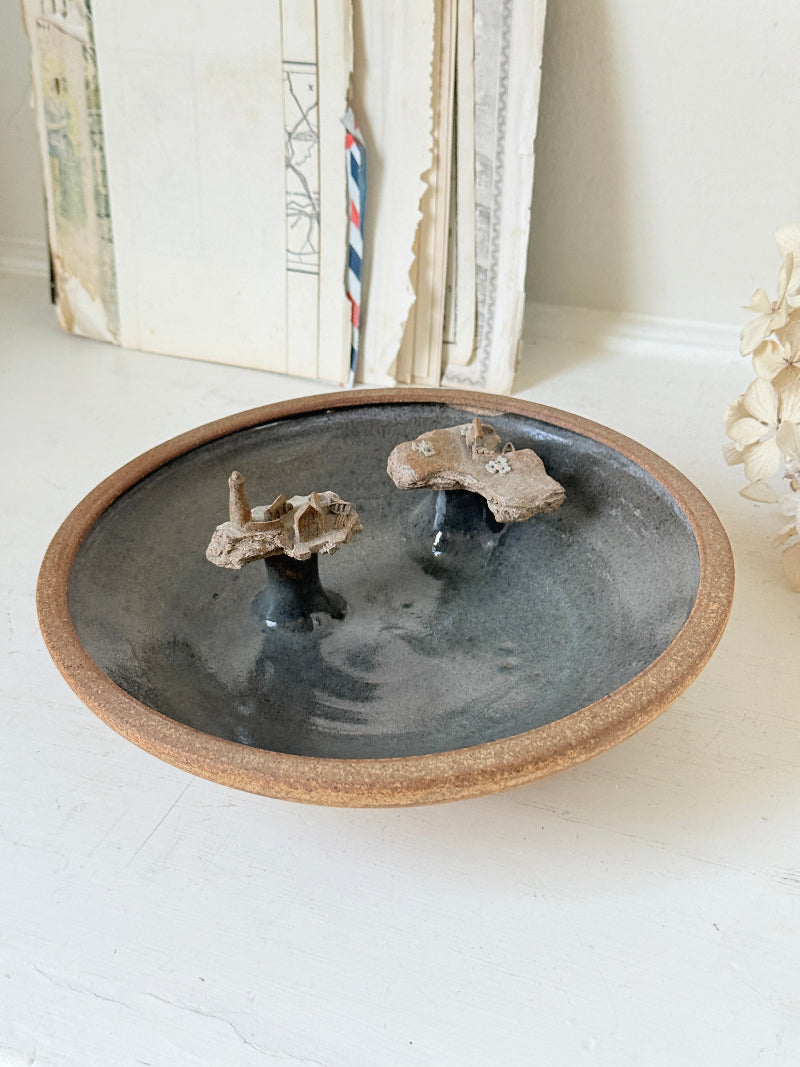 A round ceramic dish with a dark gray and black glaze, a rustic brown rim, and two small dried botanical stems placed inside, set on a white surface with books in the background.
