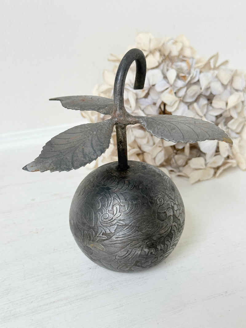 An antique-style metal hanging ornament featuring a carved round ball base with floral engravings, a curved hook top, and leaf-like wings extending from the sides, displayed against a soft white background with dried flowers behind it.