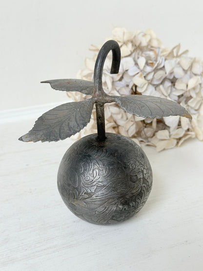An antique-style metal hanging ornament featuring a carved round ball base with floral engravings, a curved hook top, and leaf-like wings extending from the sides, displayed against a soft white background with dried flowers behind it.