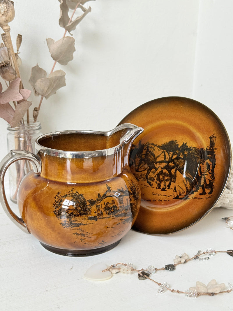 Antique Ridgways stoneware creamer jug and saucer photographed on a white surface in amber treacle glaze with black coaching scenes and silver lustre rim