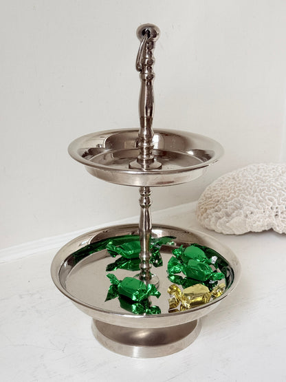 Two tier silver tone cake stand with polished reflective finish styled with green wrapped sweets on a white surface