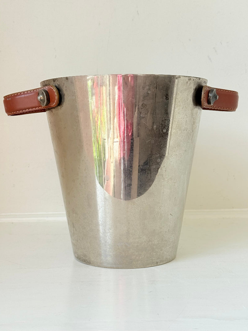 Vintage metal wine cooler with leather handles and tapered form, photographed on white surface.