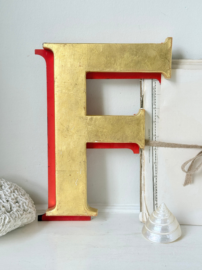 A large decorative capital letter F with a textured gold finish and red backing, styled on a white surface with a white knit fabric and a small white cone-shaped object nearby.