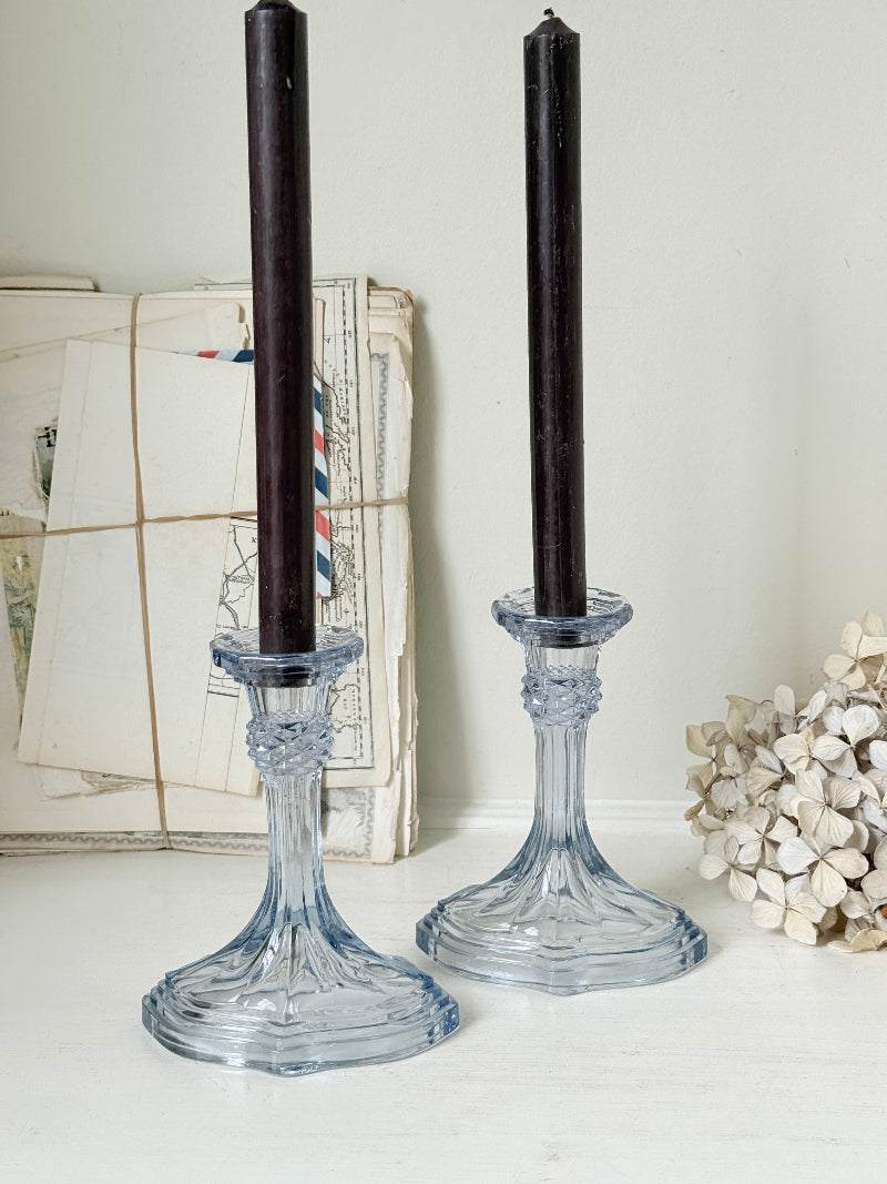 Two clear glass candlestick holders with dark taper candles, styled on a white surface with vintage papers and dried flowers in the background.