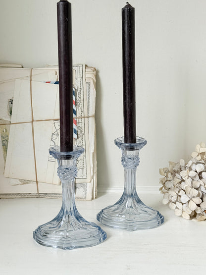 Two clear glass candlestick holders with dark taper candles, styled on a white surface with vintage papers and dried flowers in the background.