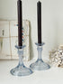 Two clear glass candlestick holders with dark taper candles, styled on a white surface with vintage papers and dried flowers in the background.