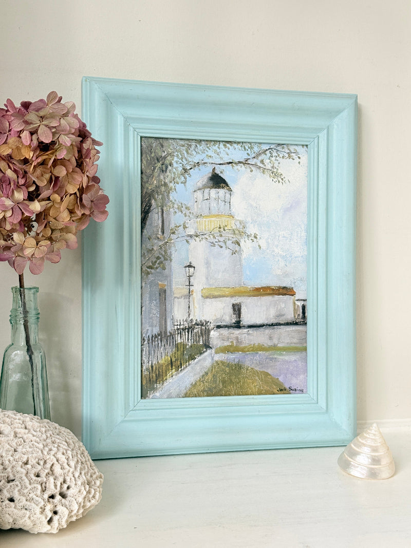A framed art print depicting a lighthouse scene, styled on a white surface next to a glass vase with pink hydrangeas, a white crochet pillow, and a small white shell. The frame is a light pastel aqua blue.