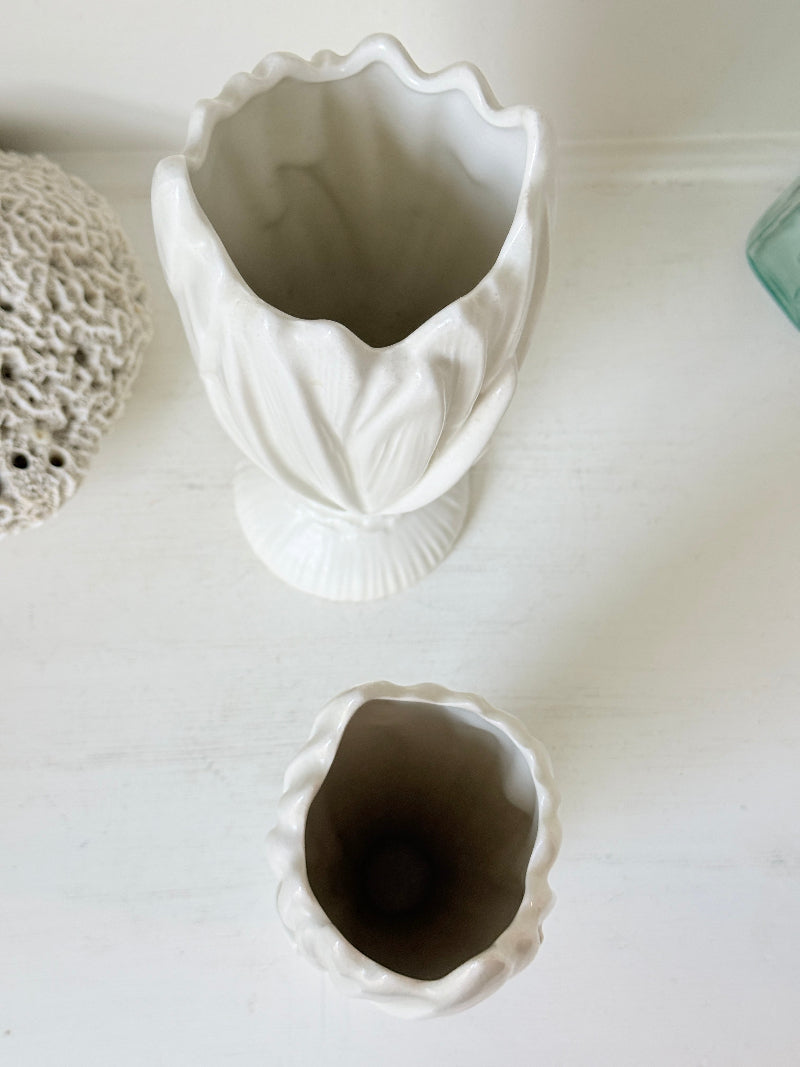 A white ceramic flower-shaped vase with a fluted rim and pedestal base, shown from above on a white surface. A smaller matching white cup or dish is placed below it, and part of a textured beige object and a blue-green glass edge are visible at the sides.