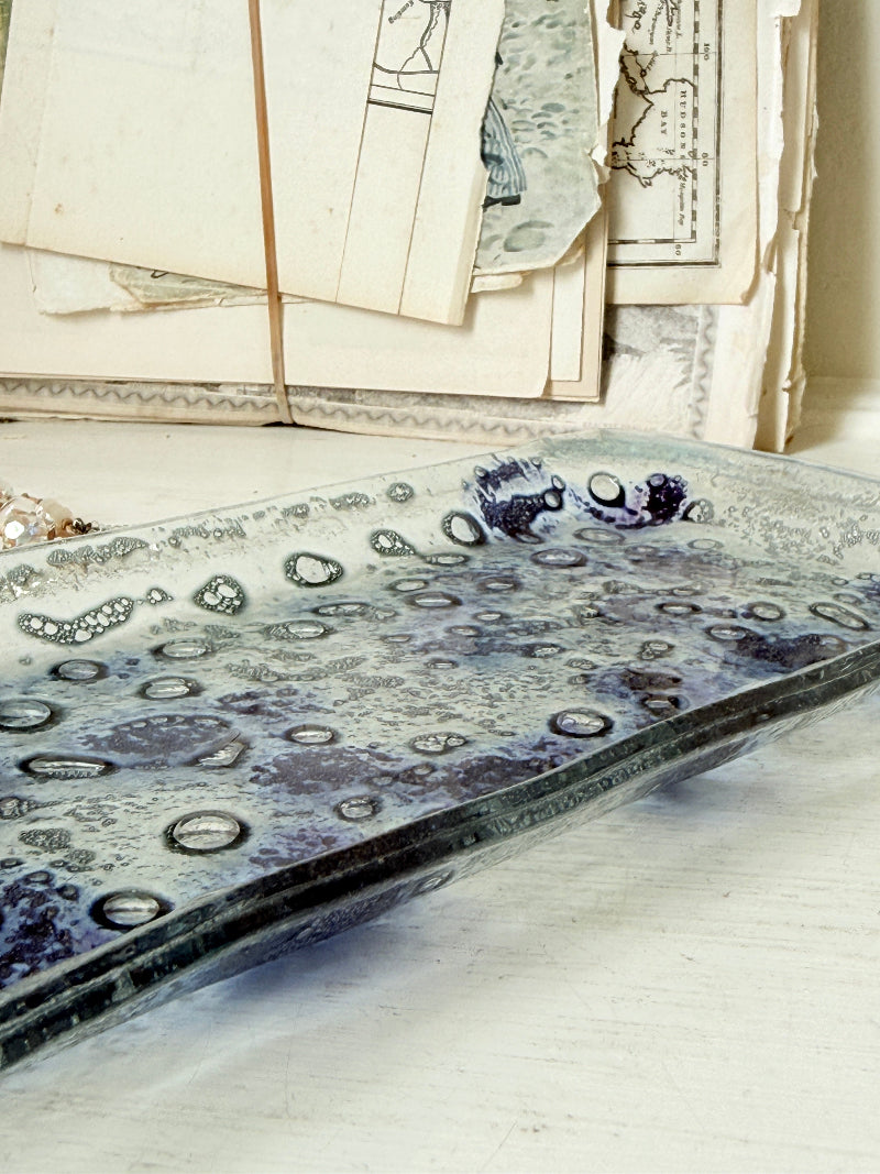 A rectangular glass serving tray with a textured, bubble-like pattern in shades of blue, gray, and white, styled on a light surface with vintage papers in the background.
