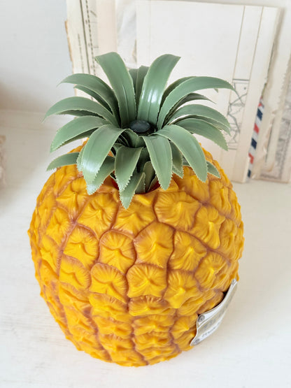 A realistic yellow pineapple-shaped decorative object with a textured scaly surface and a green leafy crown, photographed on a light tabletop with books in the background.