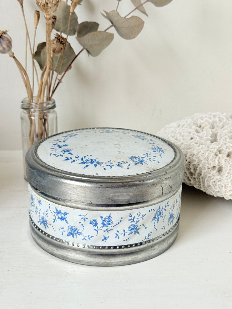 Vintage round shortbread tin with blue floral pattern on silver background, photographed on a white surface.