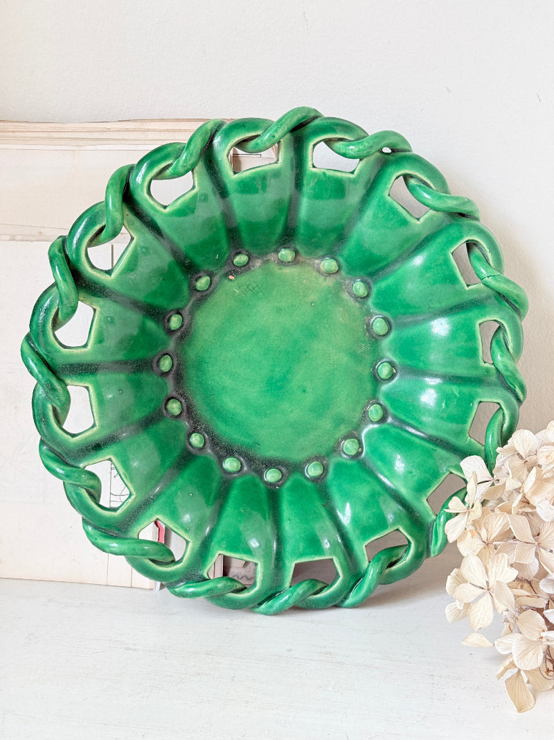 A glossy green ceramic decorative bowl with a scalloped, openwork rim and raised center, photographed against a light neutral background with dried flowers at the side.