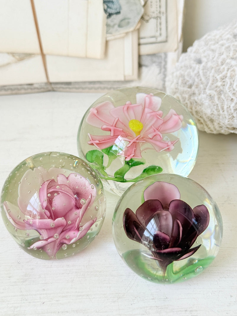 Trio of vintage glass paperweights with hand worked pink and plum floral detail, clear glass domes and green leaf decoration