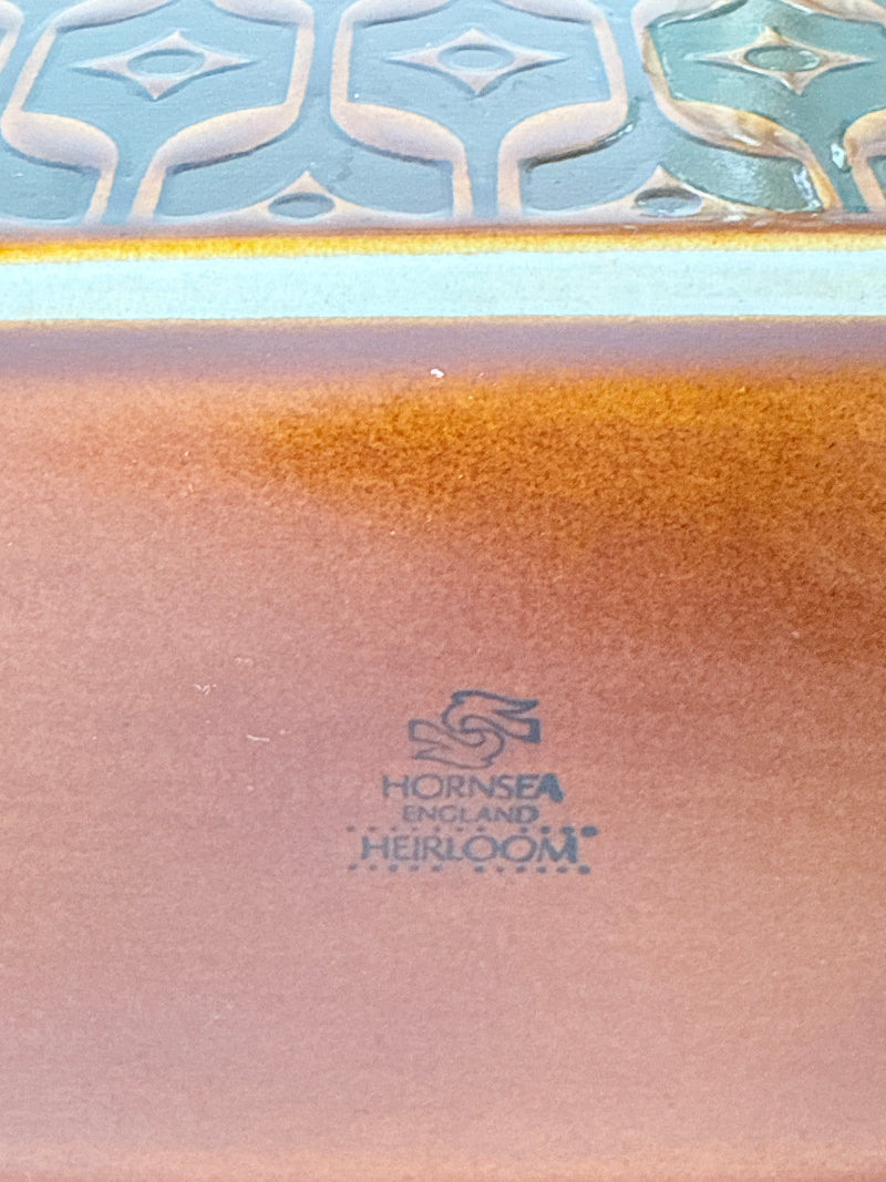 Close-up of a ceramic tile showing a glossy coppery-brown body with a blue and white patterned border and a visible Hornsea England Heirloom logo.