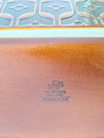 Close-up of a ceramic tile showing a glossy coppery-brown body with a blue and white patterned border and a visible Hornsea England Heirloom logo.