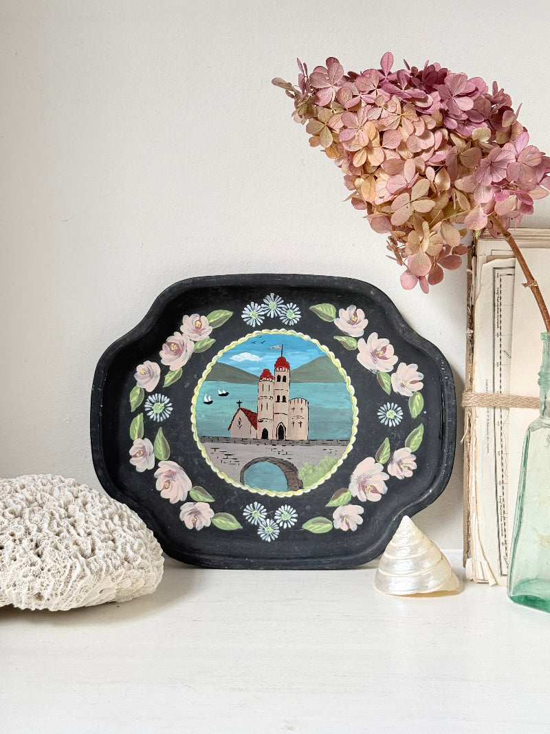 A decorative black oval tray with scalloped edges, featuring a hand-painted central scene of a castle by the water surrounded by pink flowers and green leaves. The tray is styled on a white surface with dried pink hydrangeas, a white lace cloth, a small shell, and a green glass bottle against a white wall.