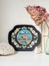 A decorative black oval tray with scalloped edges, featuring a hand-painted central scene of a castle by the water surrounded by pink flowers and green leaves. The tray is styled on a white surface with dried pink hydrangeas, a white lace cloth, a small shell, and a green glass bottle against a white wall.