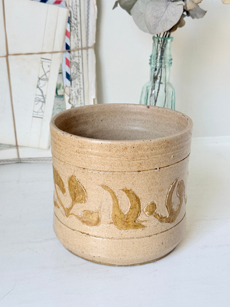 A handmade ceramic planter in a sandy beige stoneware finish with a glossy gold leaf pattern, set against a softly blurred background of vintage mail and a glass bottle vase.
